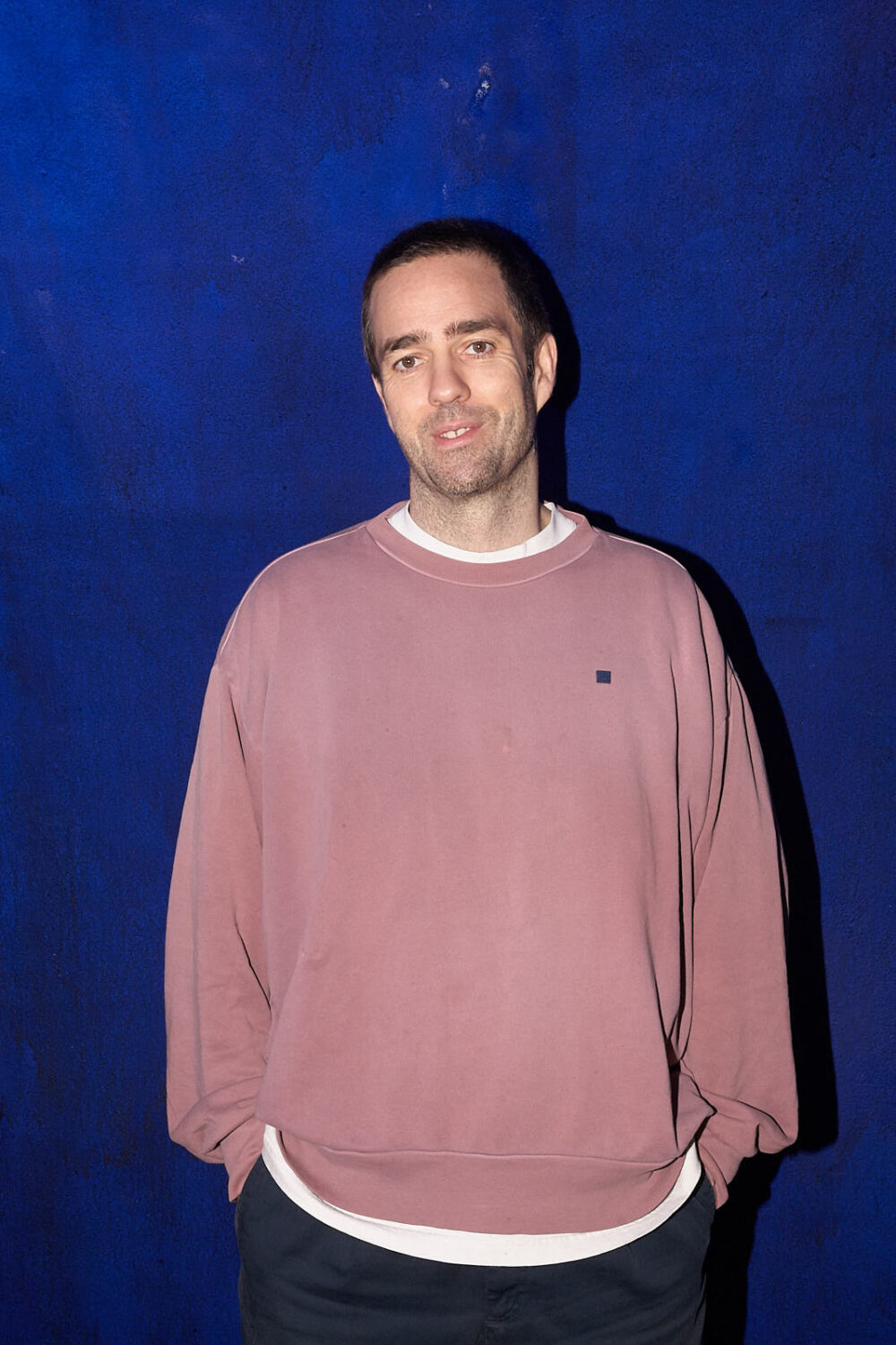 Jonathan Alphandery stands in front of a blue wall in Planque, with a faded pink sweatshirt on over a white t-shirt, with his hands in his pockets 